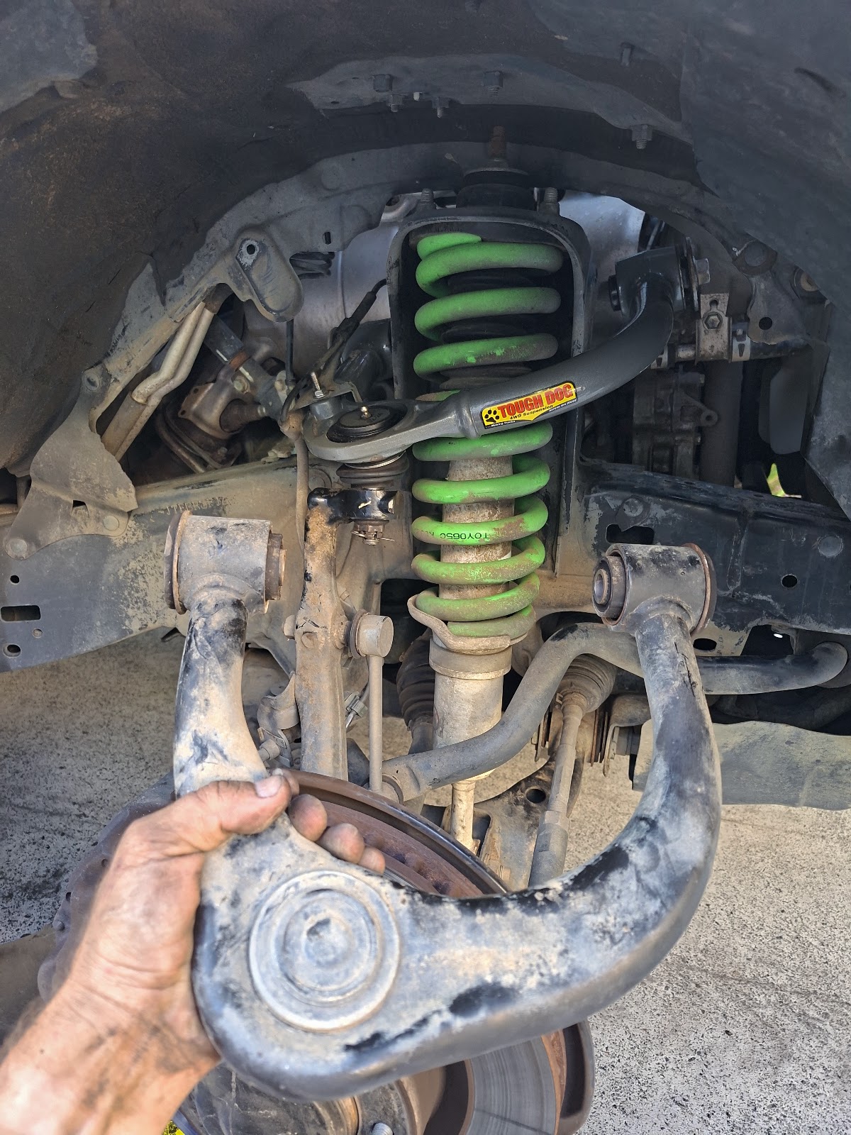 Hand holding a worn upper control arm in front of a lifted green coil spring on a 4WD