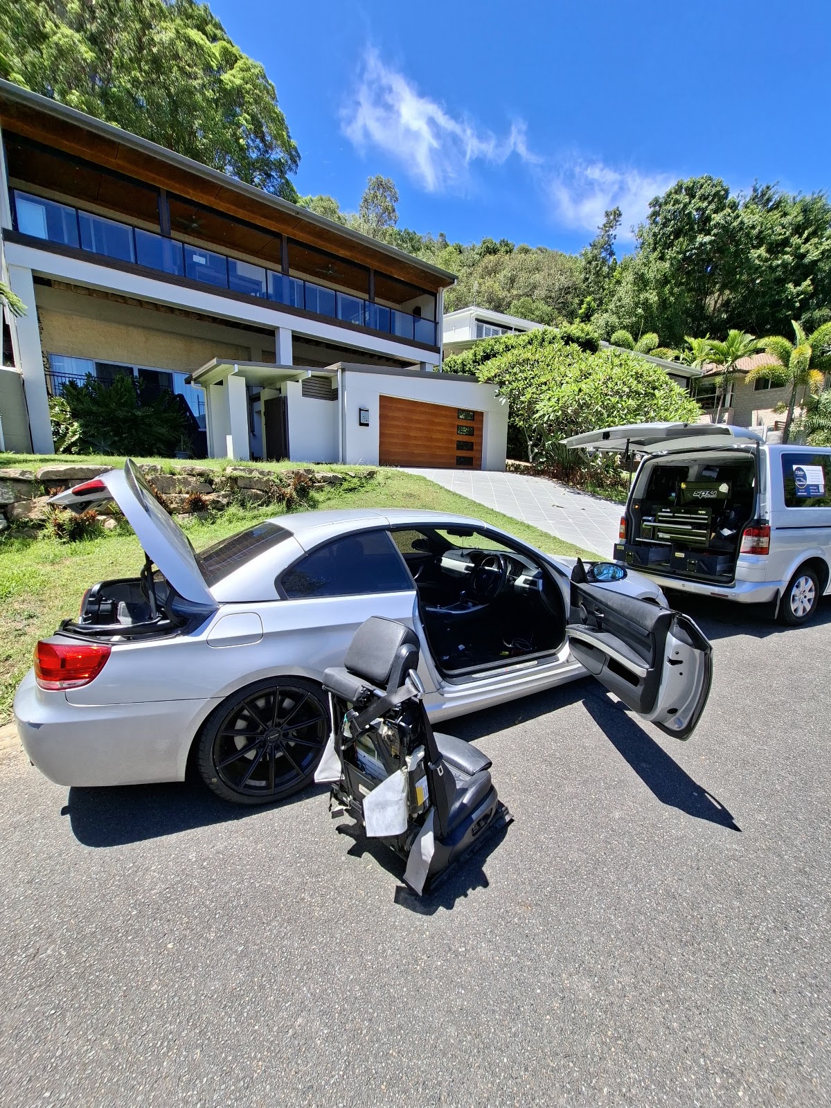 A silver BMW convertible being worked on in a hillside driveway, the L'Atelier van parked behind it